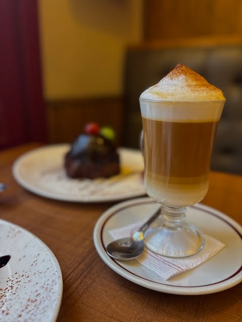 Capuchino alto con espuma y un postre de chocolate al fondo