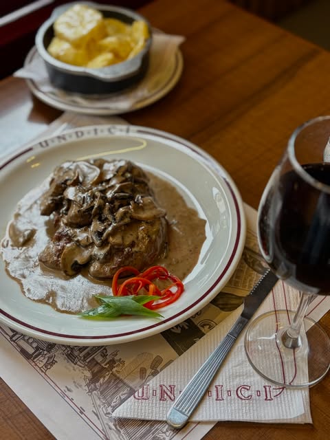 Carne con salsa de hongos y papas, acompañada de una copa de vino tinto