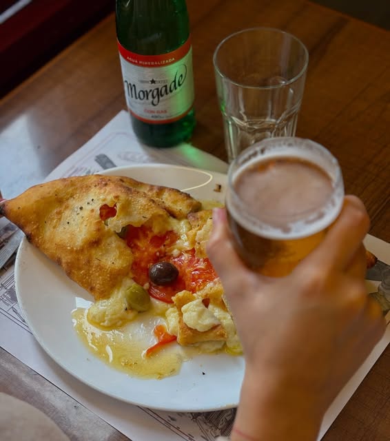 Porción de pizza con tomate y aceitunas junto a un vaso de cerveza
